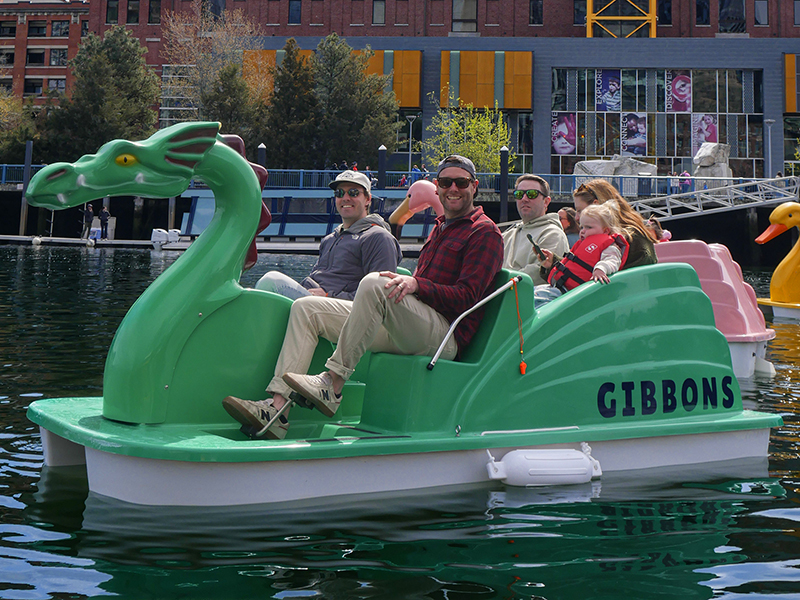 Smiling guests ride a pedal boat shaped like a dragon on the Charles River